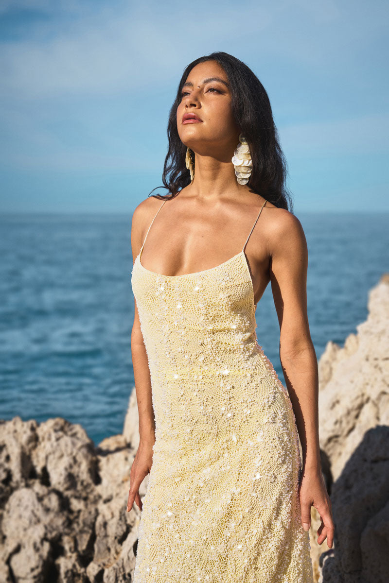 Robe Phème Paris : une robe de soirée jaune pâle à sequins scintillants et fines bretelles, issue de la nouvelle collection. Mode luxueuse conçue à Paris, présentée ici dans un décor côtier élégant pour un style resort chic unique.